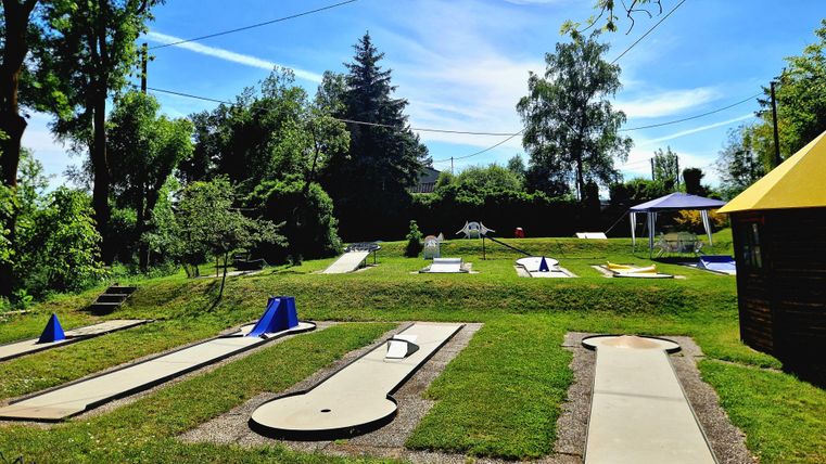 Bunte Minigolfanlage mit 18 Bahnen und grüner Rasenfläche. Im Hintergrund sind Bäume und ein blauer Himmel zu sehen. Sitzmöglichkeiten vorhanden, teilweise unter schattenspendenden Pavillons oder Baumkronen.