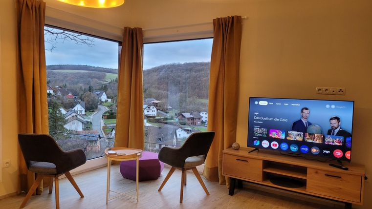 Ein gemütlicher Wohnraum mit zwei Stühlen und einem Tisch vor einem großen Fenster. Draußen ist eine ländliche Aussicht mit Hügeln und Häusern zu sehen.