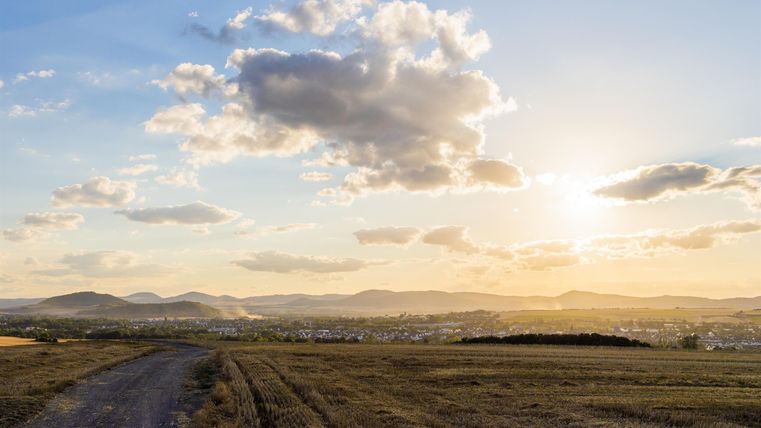 Eine malerische Landschaft mit sanften Hügeln und einem klaren Himmel. Die Sonne geht im Hintergrund unter und wirft warmes Licht über die Felder.