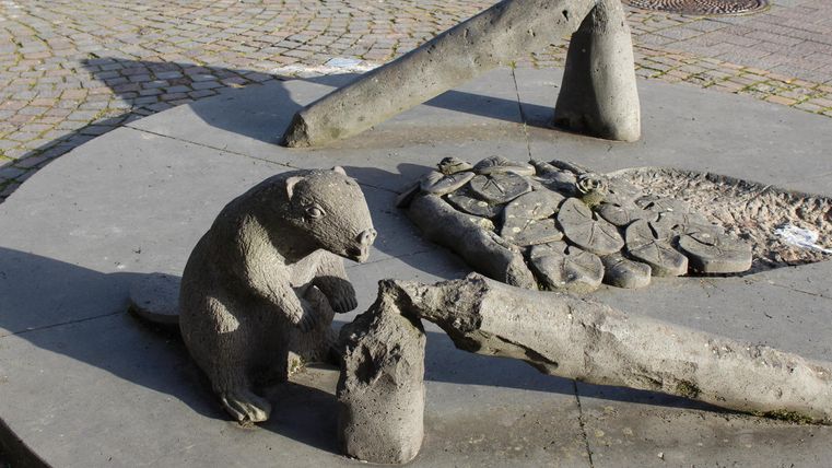 Eine kleine Skulptur eines Bären sitzt neben einem Stein- und Holzmotiv. Die Szene ist von einer gepflasterten Fläche umgeben.