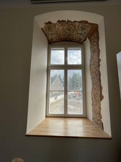 Ein Fenster mit einem Holzbrett und dekorativem Rahmen. Im Hintergrund sind Bäume und Baustellen sichtbar.