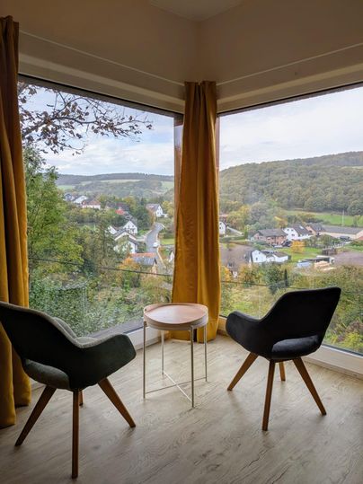 Ein gemütlicher Raum mit zwei Sesseln und einem kleinen Tisch. Durch große Fenster bietet sich ein Blick auf eine malerische Landschaft und Hügel.