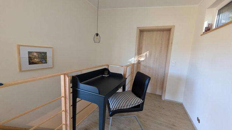 Ein heller Arbeitsbereich mit einem schwarzen Schreibtisch und einem Stuhl. Die Wände sind in einem neutralen Farbton gestrichen, und ein Fenster sorgt für Tageslicht.