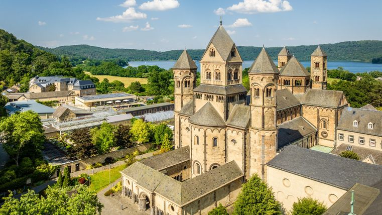 Eine beeindruckende Abtei mit einer gotischen Architektur, umgeben von grünen Bäumen. Im Hintergrund ist ein ruhiger See und ein klarer Himmel zu sehen.