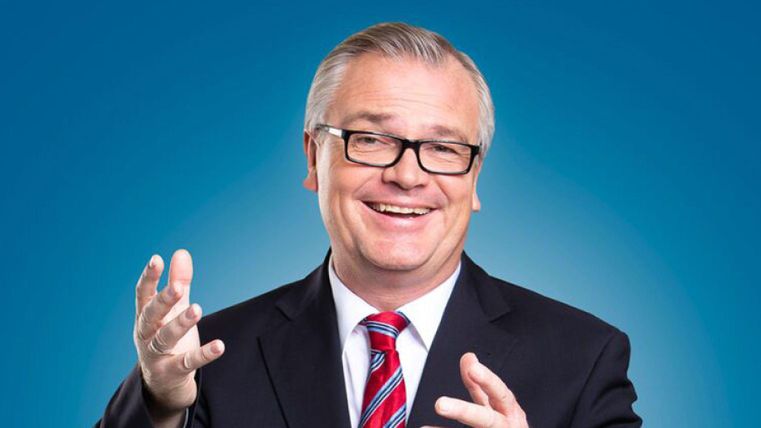 A smiling man in a suit with a tie. The background is blue and he is gesturing friendly.