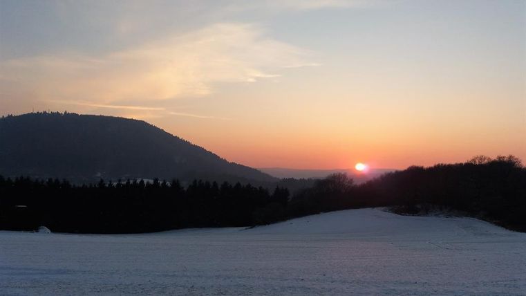 Eine malerische Winterlandschaft mit schneebedecktem Feld und einem wunderschönen Sonnenuntergang hinter den Bergen. Der Himmel zeigt sanfte Farben von Orange und Blau.