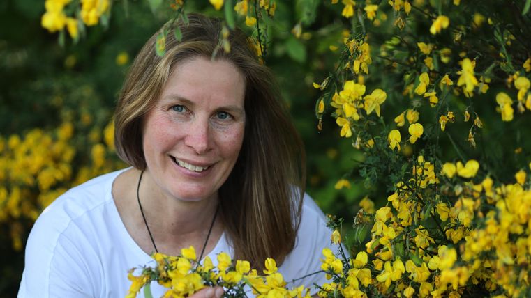 A woman smiles and stands in front of a blooming yellow plant. She holds a branch with yellow flowers in her hand.