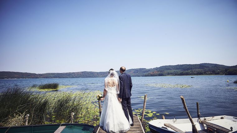Ein frisch verheiratetes Paar steht auf einem Steg am Wasser und blickt auf die Landschaft. Im Hintergrund sind Boote und eine ruhige Seenlandschaft zu sehen.