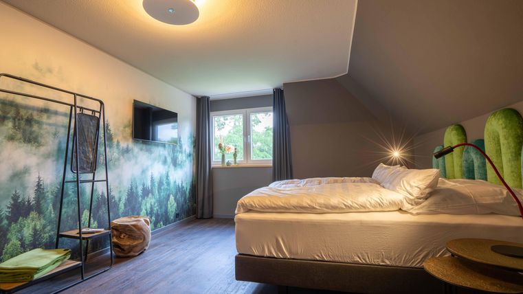 Ein modernes Schlafzimmer mit einem großen Bett und einem schönen Waldmotiv an der Wand. Es gibt ein Fenster mit Vorhängen und eine Fernseher an der Wand.