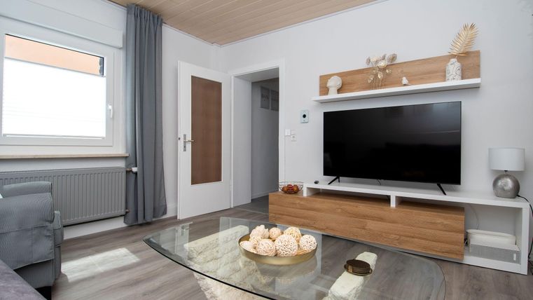 Ein modernes Wohnzimmer mit einem großen Fernseher und einem eleganten Couchtisch. Die Wände sind hell, und ein Fenster sorgt für viel natürliches Licht.