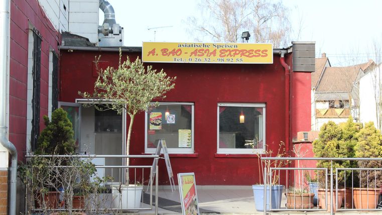A small restaurant with a red facade and a yellow sign. Several plants are on the terrace.