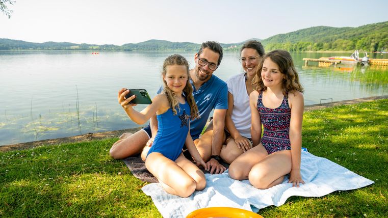 Eine Familie sitzt auf einer Decke am Ufer eines Sees. Die Kinder machen ein Selfie, während es sonnig und hell ist.