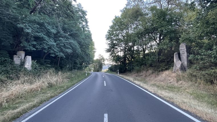 Eine ruhige Landstraße, umgeben von Bäumen und hohem Gras. Auf beiden Seiten der Straße stehen große Steinskulpturen.