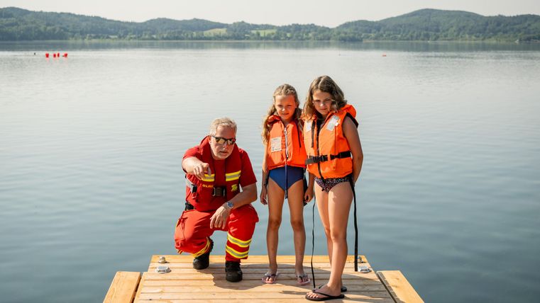 A lifeguard is standing on the dock giving instructions to two children in life jackets. In the background, a calm lake and green hills can be seen.