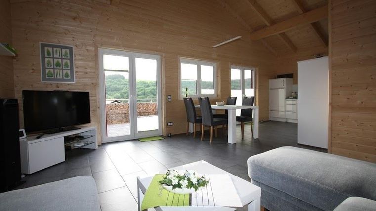 Ein modernes, helles Wohnzimmer mit Holzdecke und großen Fenstern. Es ist mit einem Fernseher, einem Esstisch und komfortablen Sofas eingerichtet.