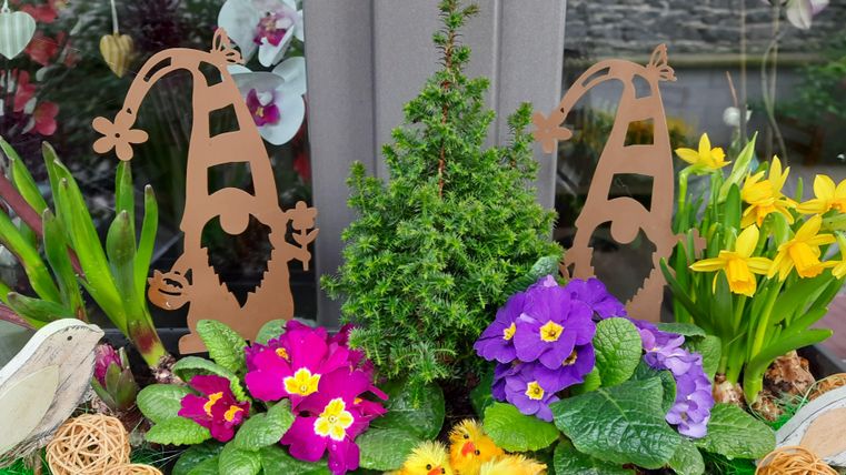 Ein bunter Blumenschmuck mit verschiedenen Pflanzen und bunten Blüten. Zwei kreative Figuren in Form von Zwergen verleihen dem Arrangement einen fröhlichen Akzent.