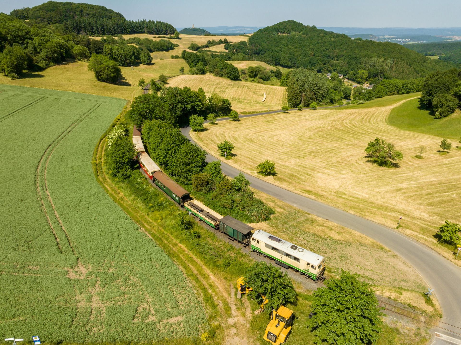 A train travels through a picturesque landscape with gentle hills and lush greenery. The fields are well-kept, and there is a bright blue sky visible.