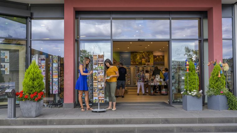 Twee vrouwen staan voor een winkel en bekijken ansichtkaarten. Op de achtergrond zijn meer klanten in de winkel te zien.