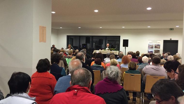 Eine große Gruppe von Menschen sitzt in einem Raum und hört einem Vortrag zu. Im Hintergrund sind einige Redner und eine Bühne sichtbar.