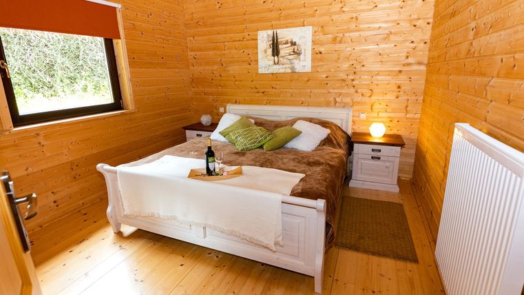 A cozy bedroom with wooden-paneled walls. The bed is adorned with soft pillows and a blanket, and on the nightstand sits a bottle of wine.