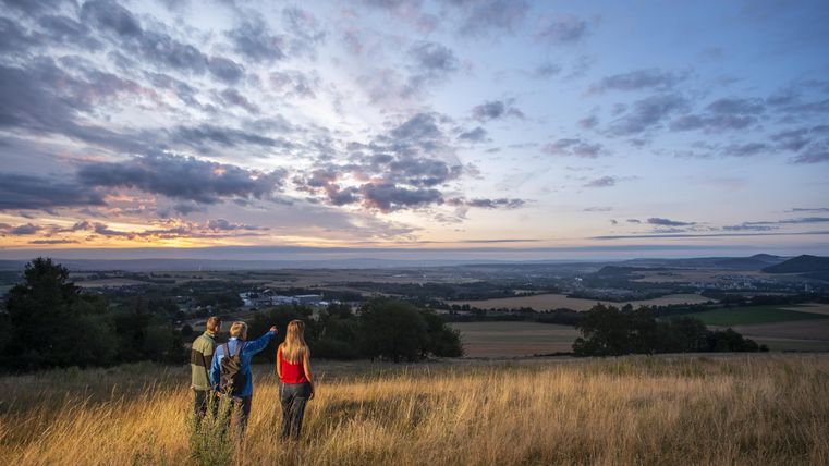 Drei Personen stehen auf einem Hügel und schauen auf die weite Landschaft. Der Himmel ist mit Wolken gefüllt und es ist kurz vor Sonnenuntergang.