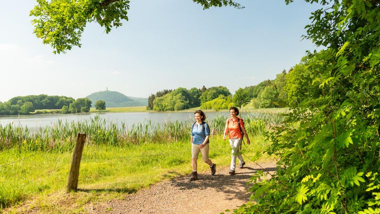 Zwei Frauen gehen entlang eines Wanderwegs an einem ruhigen See. Im Hintergrund sind grüne Wälder und ein sanfter Hügel zu sehen.