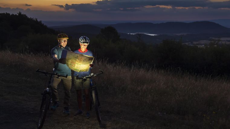 Zwei Radfahrer stehen bei Sonnenuntergang auf einem Weg und schauen auf eine Karte. Die Landschaft im Hintergrund zeigt sanfte Hügel und einen See.