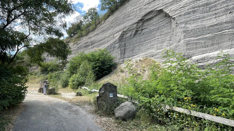 Ein Weg führt entlang einer steilen Felswand, umgeben von Bäumen und Sträuchern. Das Gebiet wirkt natürlich und ruhig.