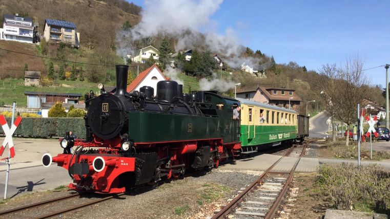 Eine Dampflokomotive fährt mit einem bunten Wagen durch eine malerische Landschaft. Im Hintergrund sind Häuser und sanfte Hügel zu sehen.