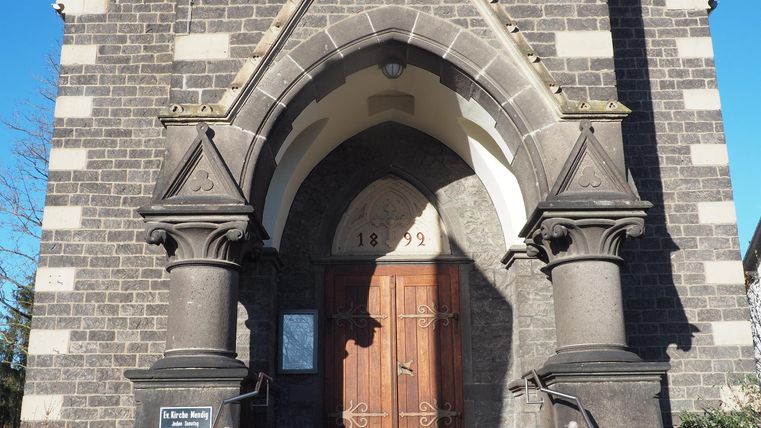 Ein historisches Gebäude mit einer markanten Fassade und großen Säulen. Die Eingangstür ist aus Holz und umgeben von einem architektonisch interessanten Bogen.