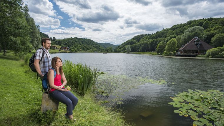 Zwei Personen sitzen am Ufer eines Sees mit bewaldeten Hügeln im Hintergrund.
