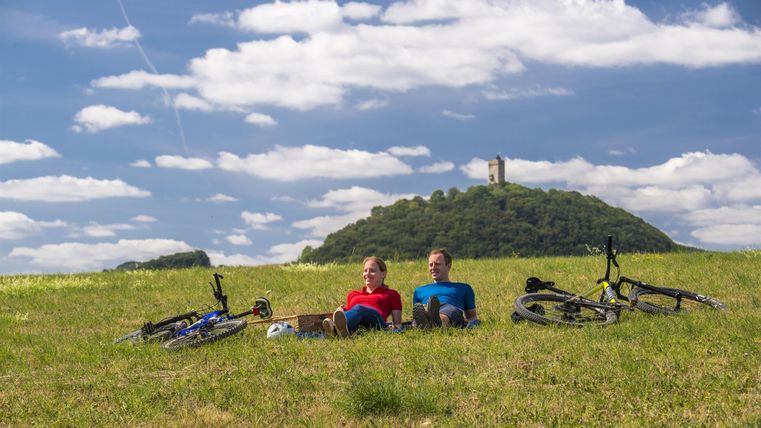 Zwei Männer liegen entspannend auf einer Wiese mit ihren Fahrrädern. Im Hintergrund ist ein Hügel mit einer Burg zu sehen, unter einem strahlend blauen Himmel.