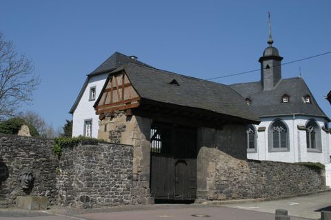 Ein historisches Gebäude mit einer Steinmauer und einem Holztor. Im Hintergrund ist eine Kirche mit einem markanten Turm sichtbar.