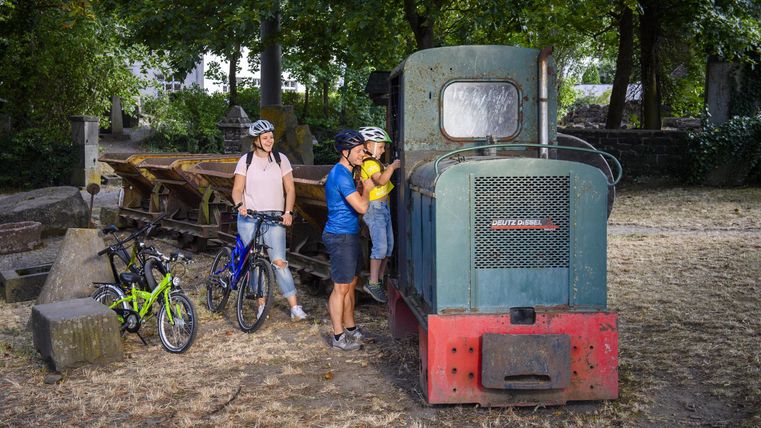 A group of cyclists stands next to a green locomotive. In the background, trees and a shady spot can be seen.