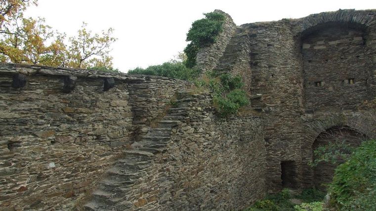 An old stone ruin with a staircase and lush vegetation. In the background, remnants of walls and a gate-like entrance are visible.