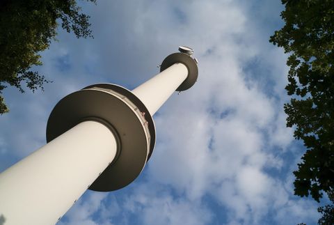 Ein hoher Aussichtsturm steht unter einem klaren, wolkigen Himmel. Umgeben von Bäumen bietet er einen eindrucksvollen Blick nach oben.