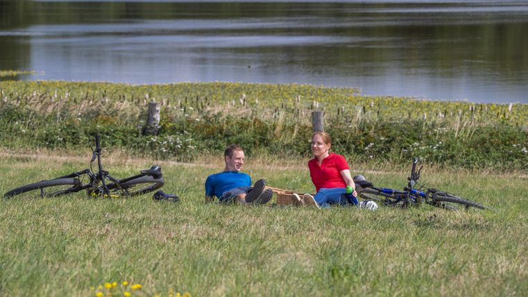 Ein Paar sitzt entspannt auf einer grünen Wiese neben einem See. Ihre Fahrräder liegen neben ihnen im Gras.