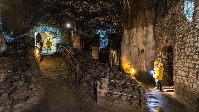 An underground cave area with stone walls and a paved path. Some people in yellow rain jackets are exploring the surroundings with flashlights.