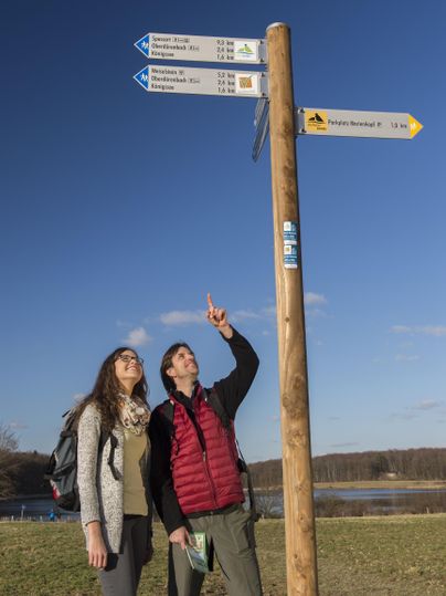 Two people are standing at a signpost outdoors. They are looking at the signs that indicate different hiking trails.