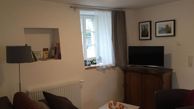 Ein gemütliches Wohnzimmer mit einem Sofa, einem Fernseher und einem Tisch. Der Raum hat ein Fenster mit Vorhängen und ist hell erleuchtet.