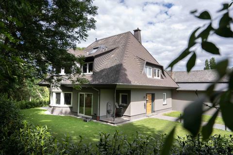 A modern house with a sloping roof and large windows. The garden is green and well-kept, surrounded by trees.
