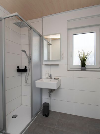 Ein modernes Badezimmer mit einer Dusche und einem kleinen Waschbecken. Helle Fliesen und ein Fenster sorgen für eine angenehme Atmosphäre.
