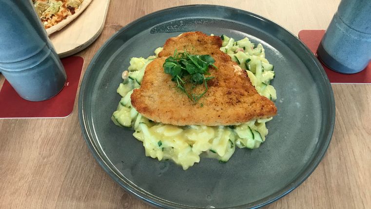 Ein leckeres Gericht mit paniertem Fisch auf einem Bett aus cremigen Kartoffeln. Dazu frische Kräuter als Garnitur auf einem grauen Teller.