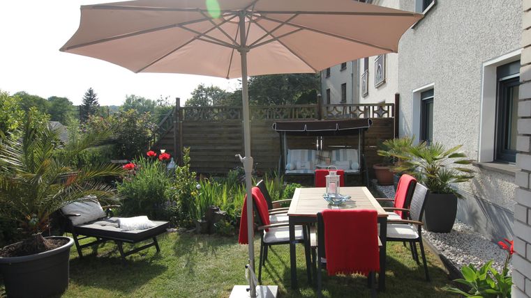 Ein einladender Garten mit einem Tisch und Stühlen unter einem großen Sonnenschirm. Im Hintergrund blühen Pflanzen und es gibt eine Liege zum Entspannen.