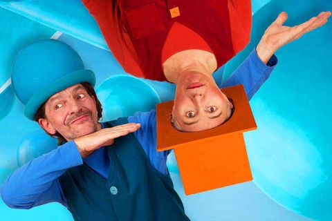 Zwei komische Figuren in bunten Outfits posieren vor einem blauen Hintergrund. Eine Figur trägt einen großen blauen Hut und die andere eine orangefarbene Mütze.
