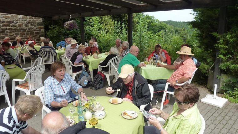 Eine Gruppe von Menschen sitzt an Tischen im Freien und genießt Essen und Getränke. Die Atmosphäre ist entspannt, umgeben von grüner Natur.