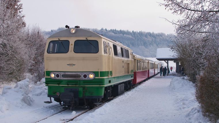 Eine verschneite Landschaft mit schneebedeckten Bäumen und Schienen, die sich durch den Wald schlängeln. Eine kleine Brücke überquert die Schienen.