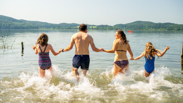 Eine Gruppe von vier Personen, darunter zwei Erwachsene und zwei Kinder, läuft lächelnd ins Wasser. Die Umgebung ist sonnig mit Bergen im Hintergrund.