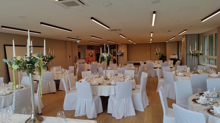 Ein elegant dekorierter Veranstaltungsraum mit runden Tischen und weißen Tischdecken. Die Tische sind mit Blumenarrangements und Kerzenständern geschmückt.