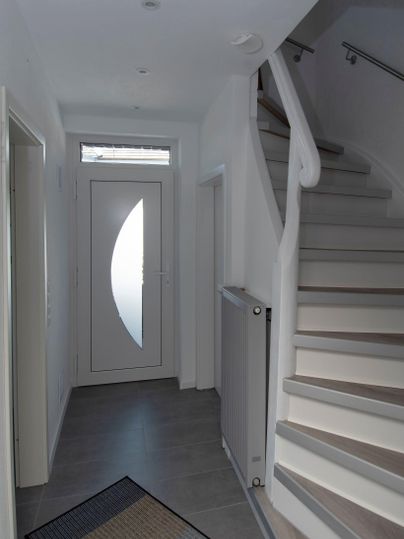 Ein heller Eingangsbereich mit einer modernen Treppe. Die Wände sind weiß und es gibt eine Tür mit einem dekorativen Fenster.